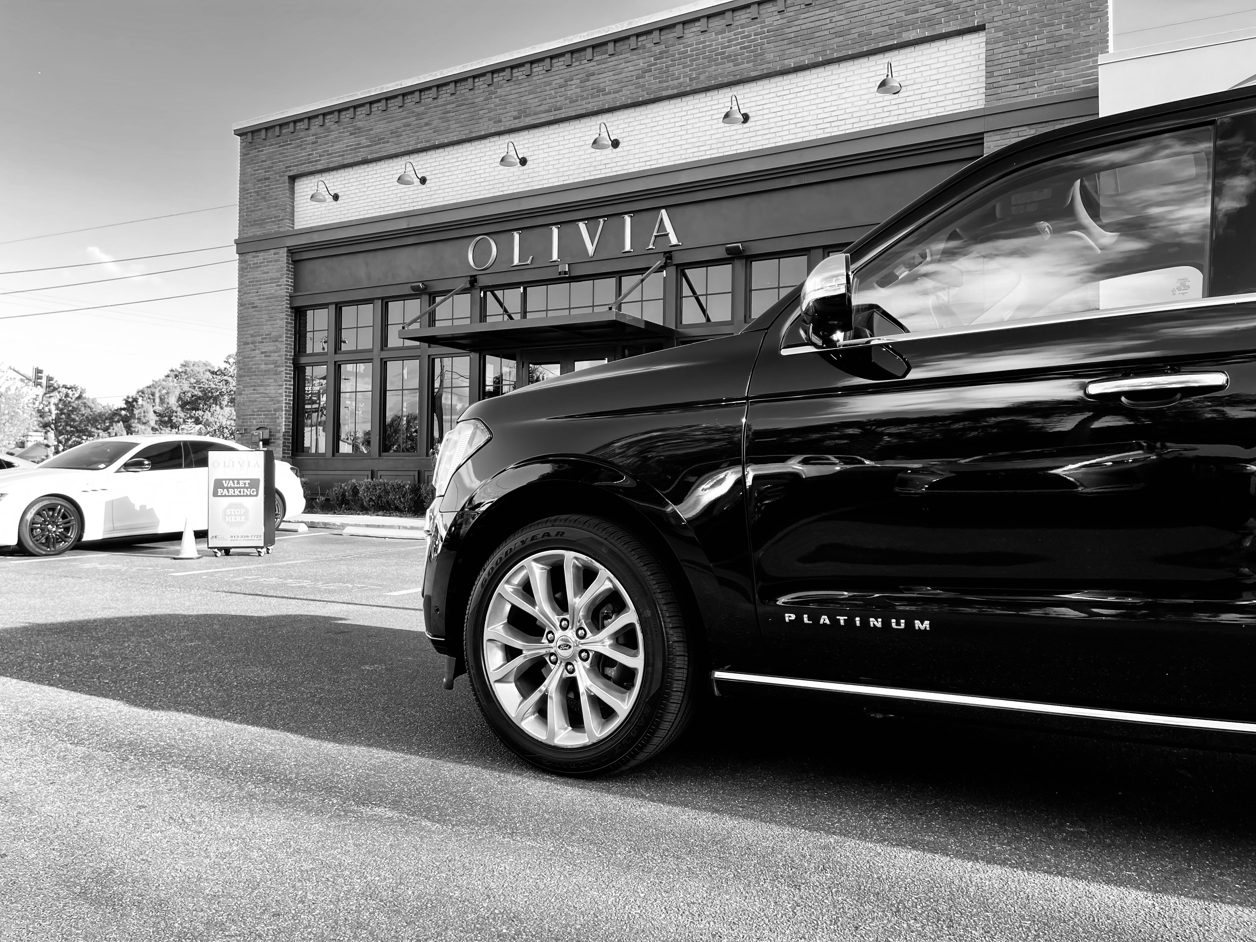 A black luxury SUV parked outside a restaurant named Olivia, with a white sports car visible in the background and a valet parking sign in front.